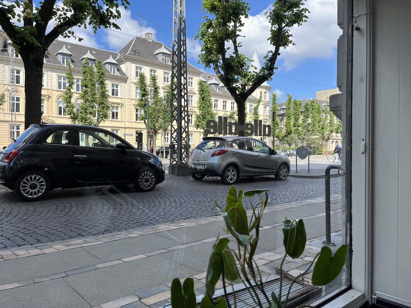 Udsigt gennem butiksruden med allé-alle-skiltet spejlvendt og to parkerede biler på Frederiksberg Allé